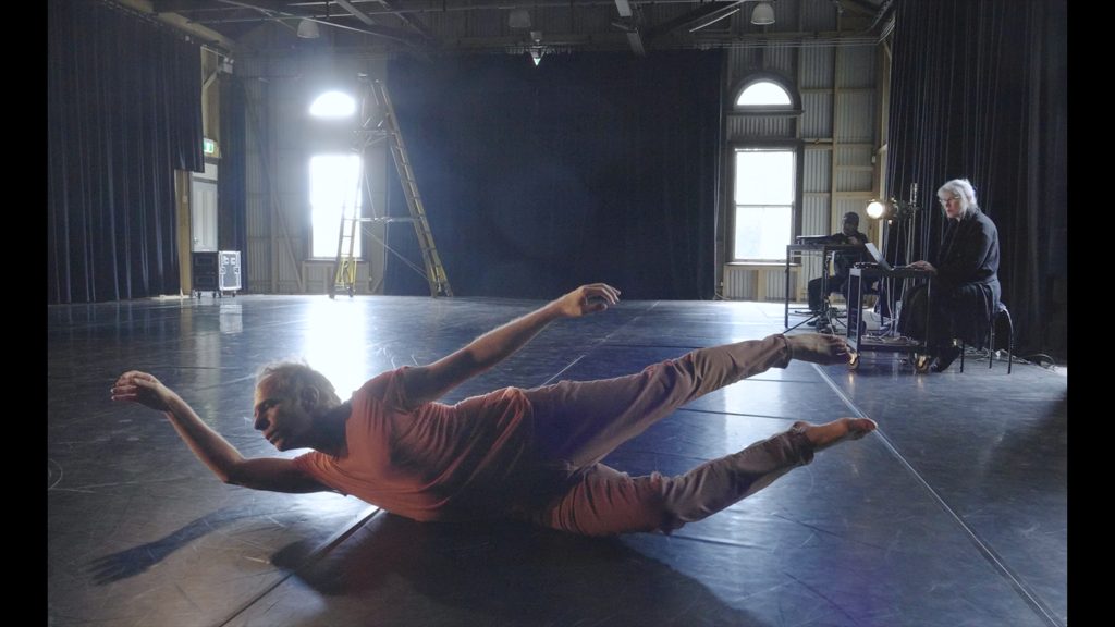 A person dancing; they are on the floor balancing on the side of their pelvis with arms, head and feet reaching up into the air.
