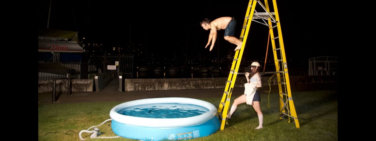 An image taken at nighttime outside The Drill Hall on lawn beside the harbour. A person stands holding onto a large yellow ladder. Another person is captured in flight diving off the ladder into an inflatable baby pool.