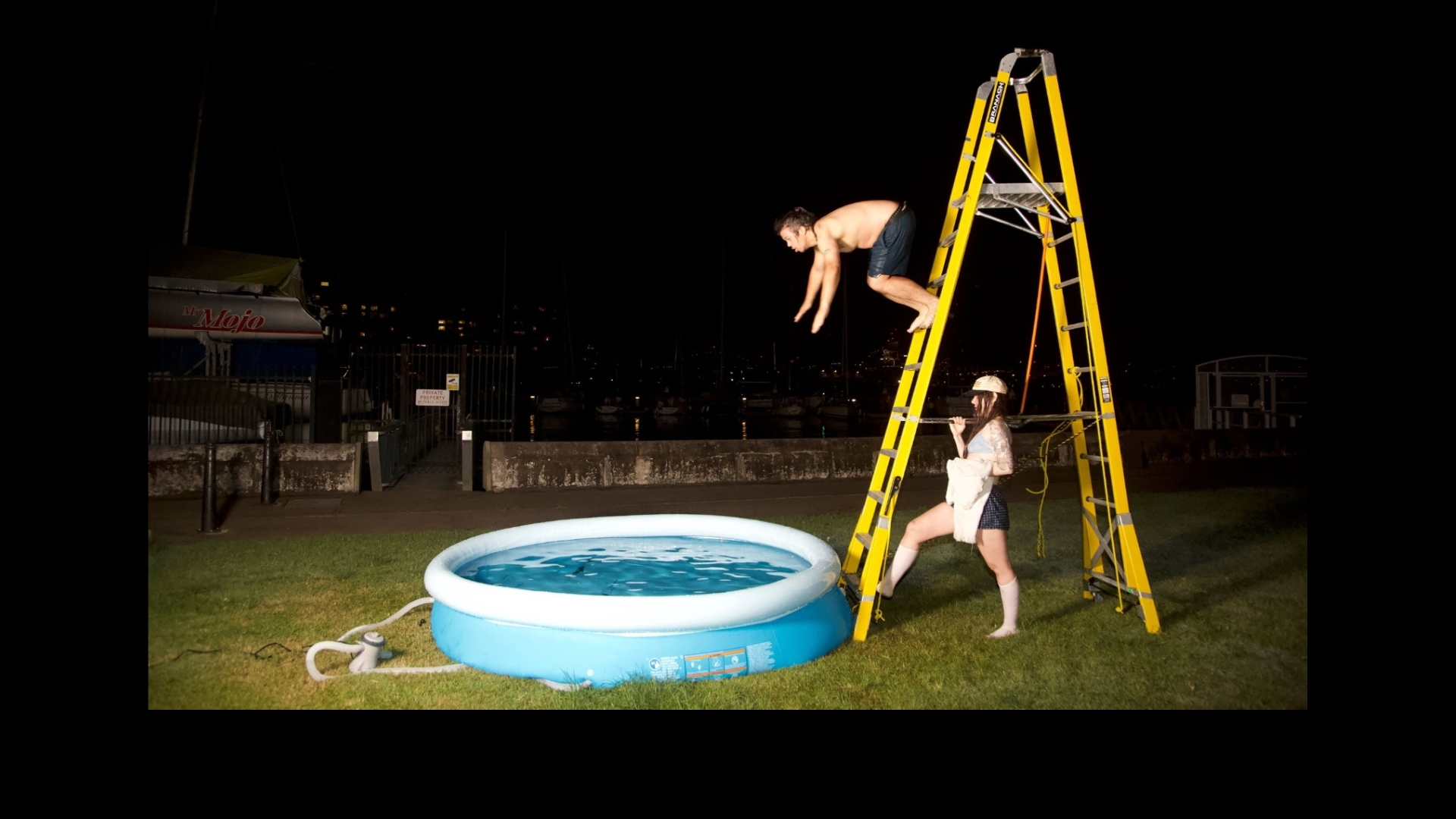 An image taken at nighttime outside The Drill Hall on lawn beside the harbour. A person stands holding onto a large yellow ladder. Another person is captured in flight diving off the ladder into an inflatable baby pool.