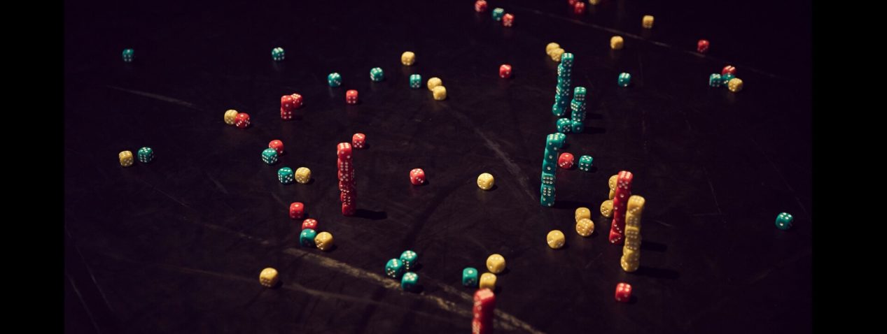 An image of coloured dice scattered on a black floor. There are many small towers of dice, each with 6 or 7 dice balancing on top of each other.