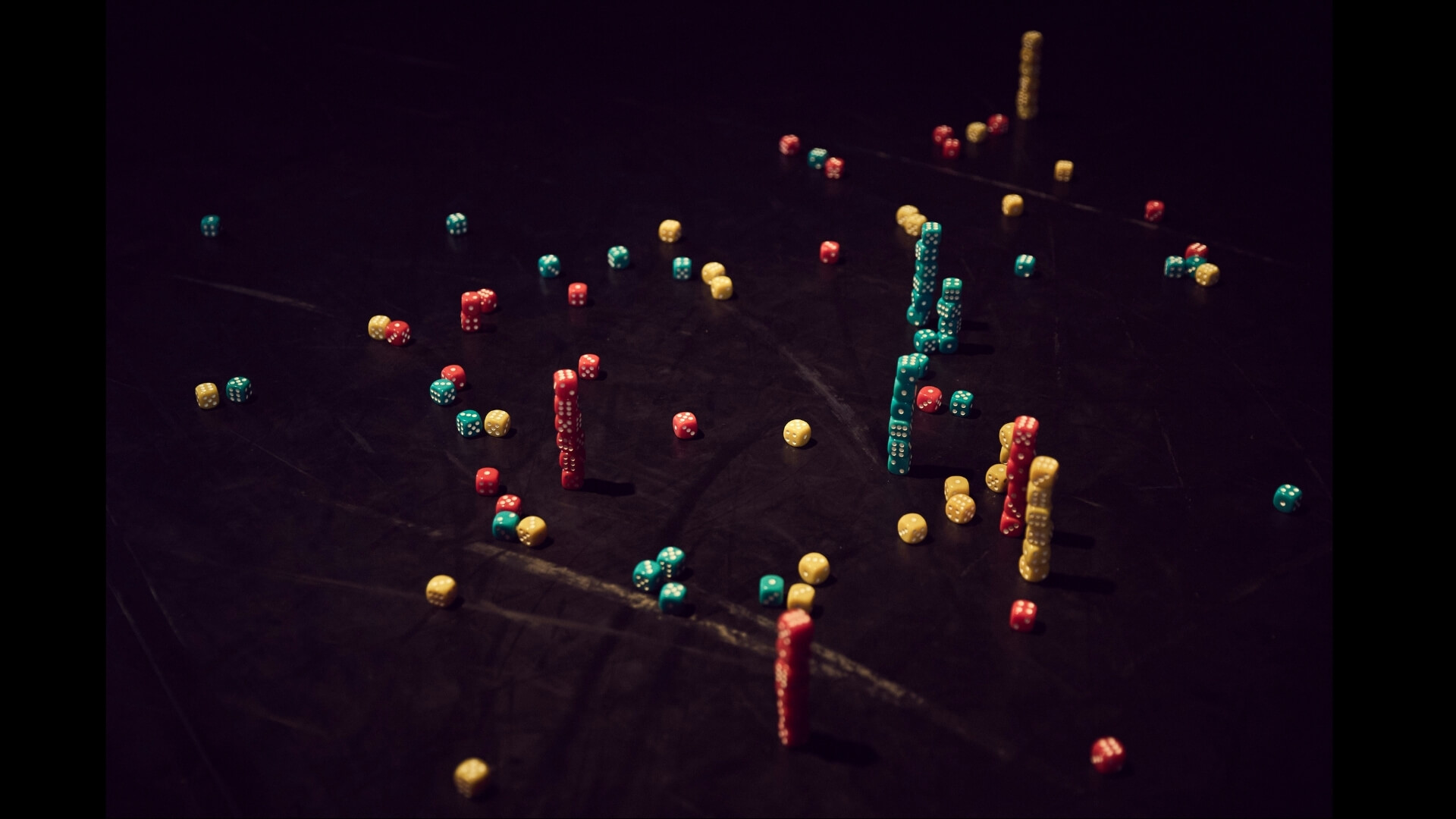 An image of coloured dice scattered on a black floor. There are many small towers of dice, each with 6 or 7 dice balancing on top of each other.