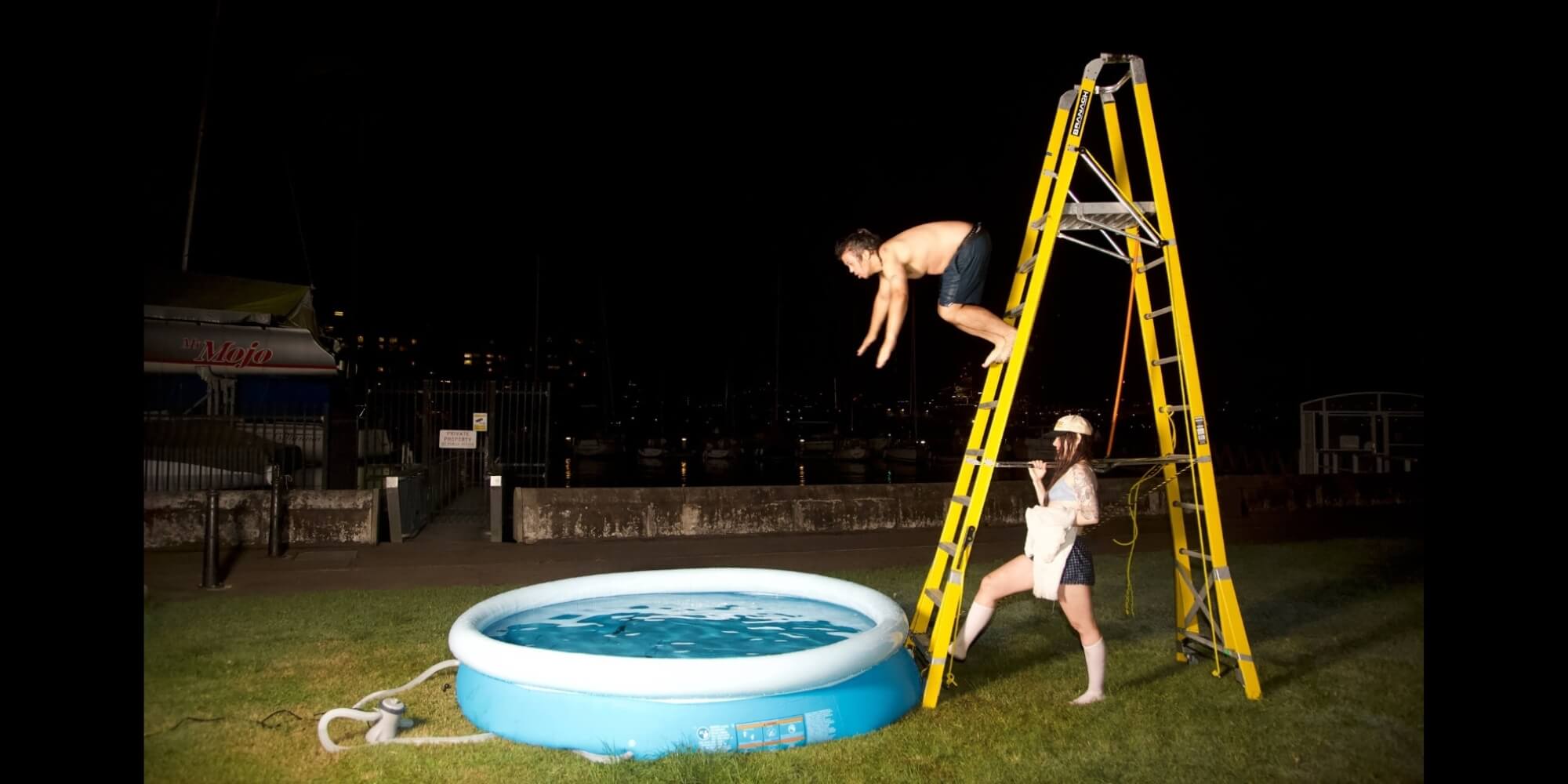 An image taken at nighttime outside The Drill Hall on lawn beside the harbour. A person stands holding onto a large yellow ladder. Another person is captured in flight diving off the ladder into an inflatable baby pool.