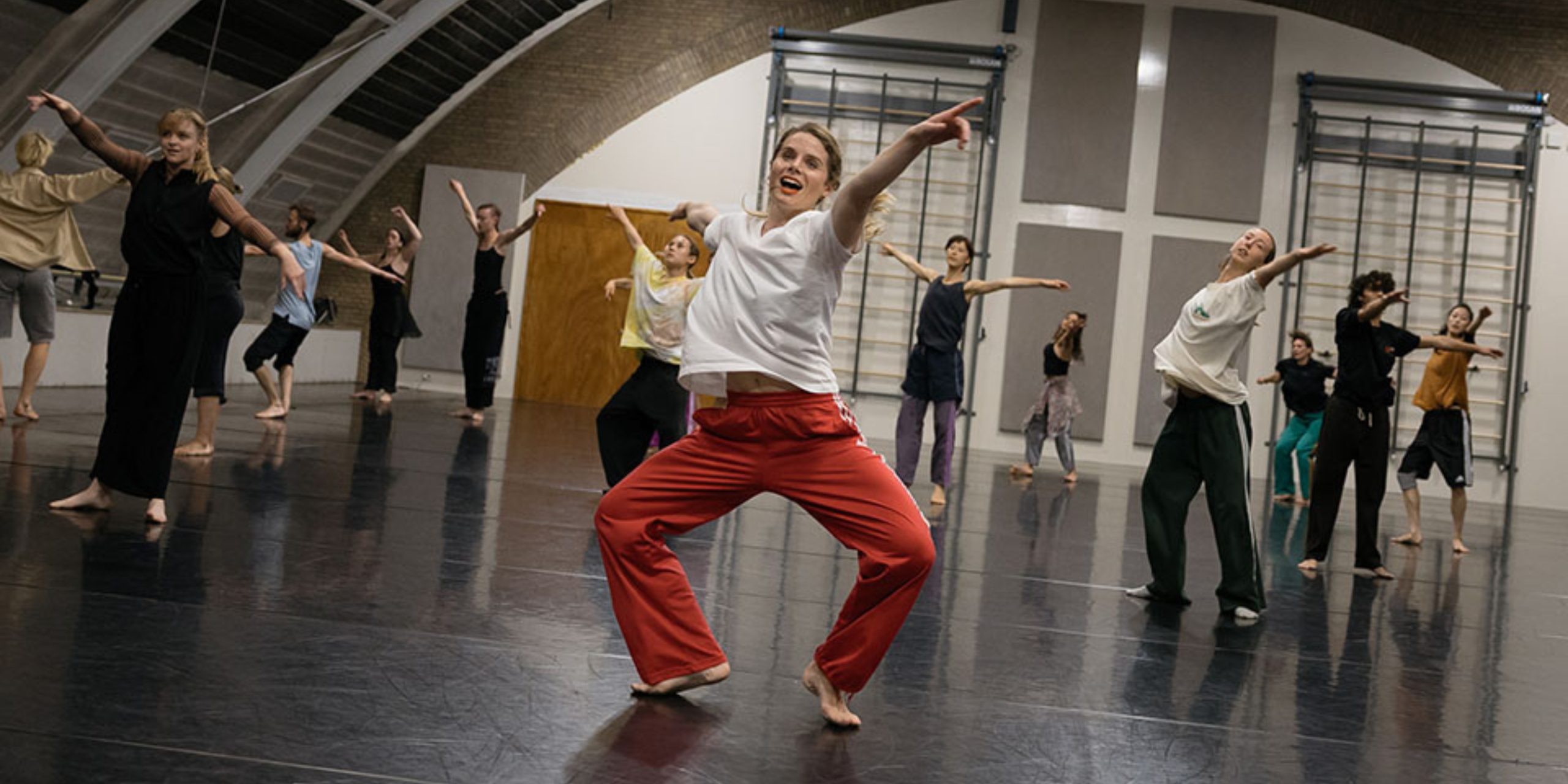 A lively group of dancers moves expressively across a large dance studio. At the centre, a dancer in pink pants and a white top leads with joyful energy, arms open wide. The other dancers follow in rhythmic, expansive gestures, filling the space with motion and connection.