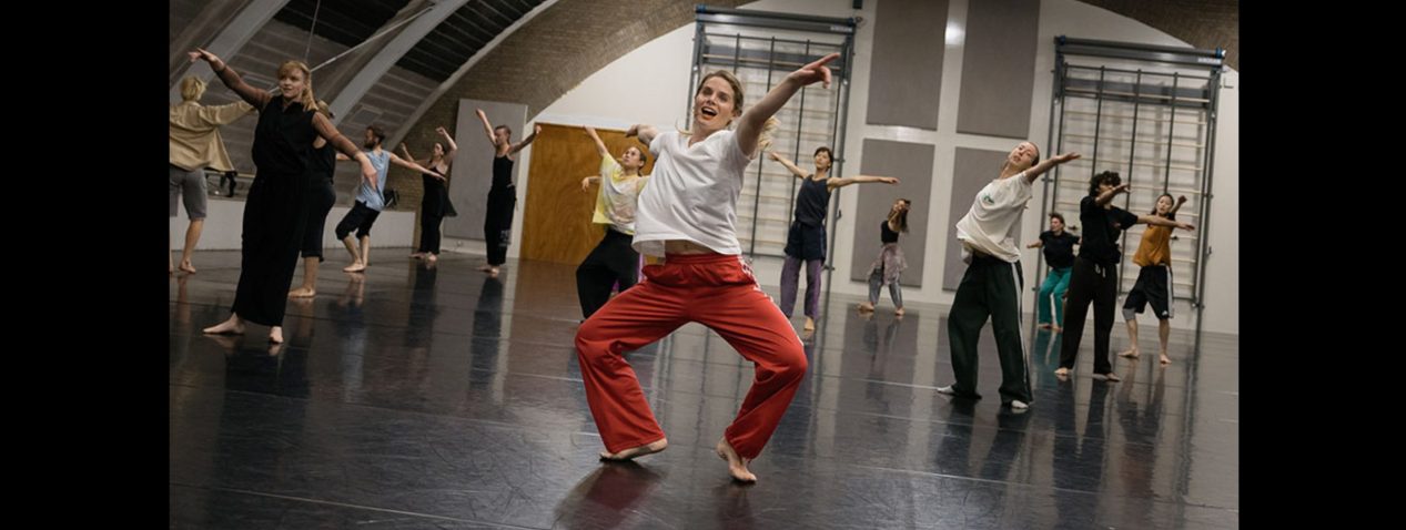 A lively group of dancers moves expressively across a large dance studio. At the centre, a dancer in pink pants and a white top leads with joyful energy, arms open wide. The other dancers follow in rhythmic, expansive gestures, filling the space with motion and connection.