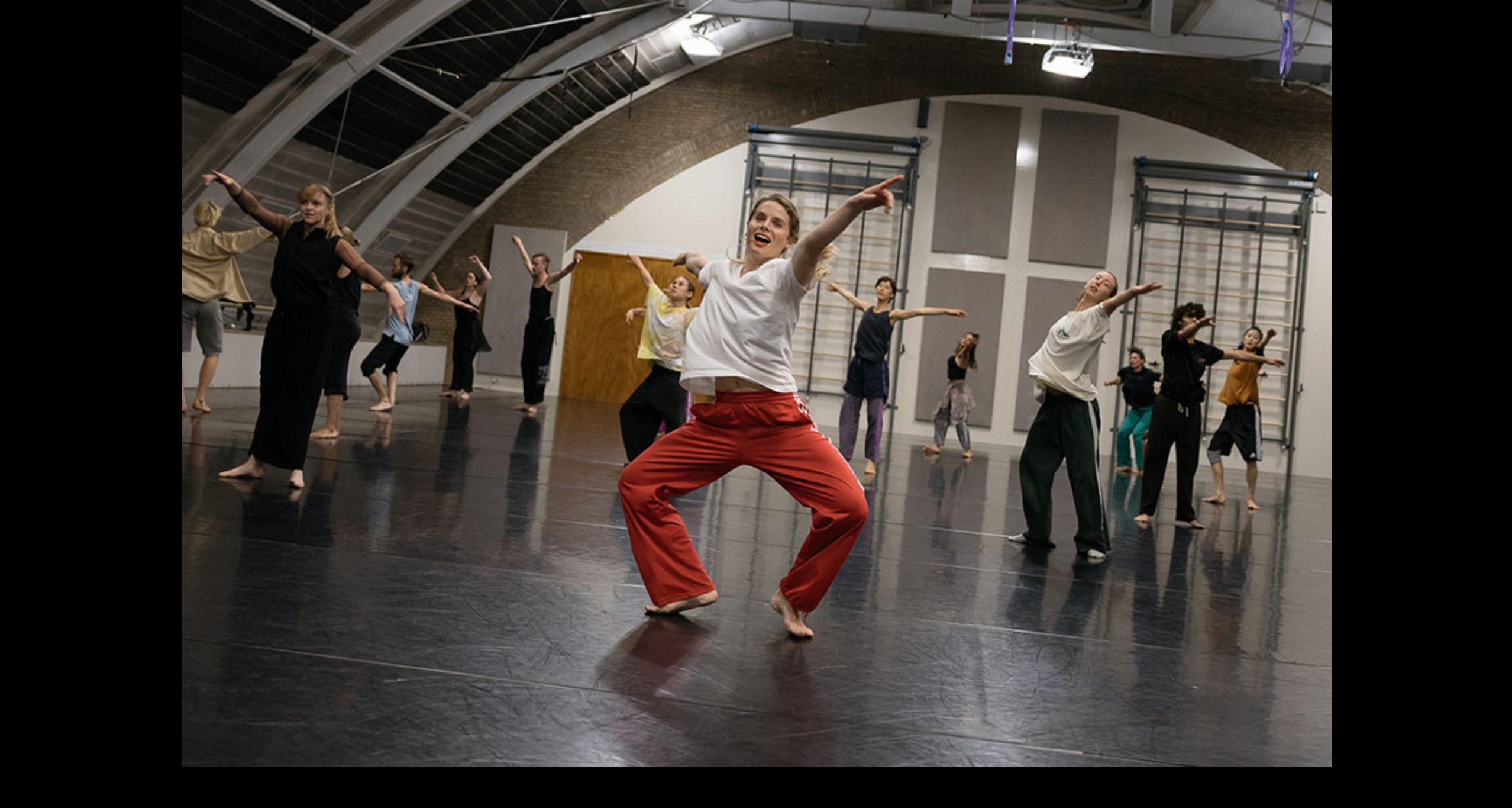 A lively group of dancers moves expressively across a large dance studio. At the centre, a dancer in pink pants and a white top leads with joyful energy, arms open wide. The other dancers follow in rhythmic, expansive gestures, filling the space with motion and connection.
