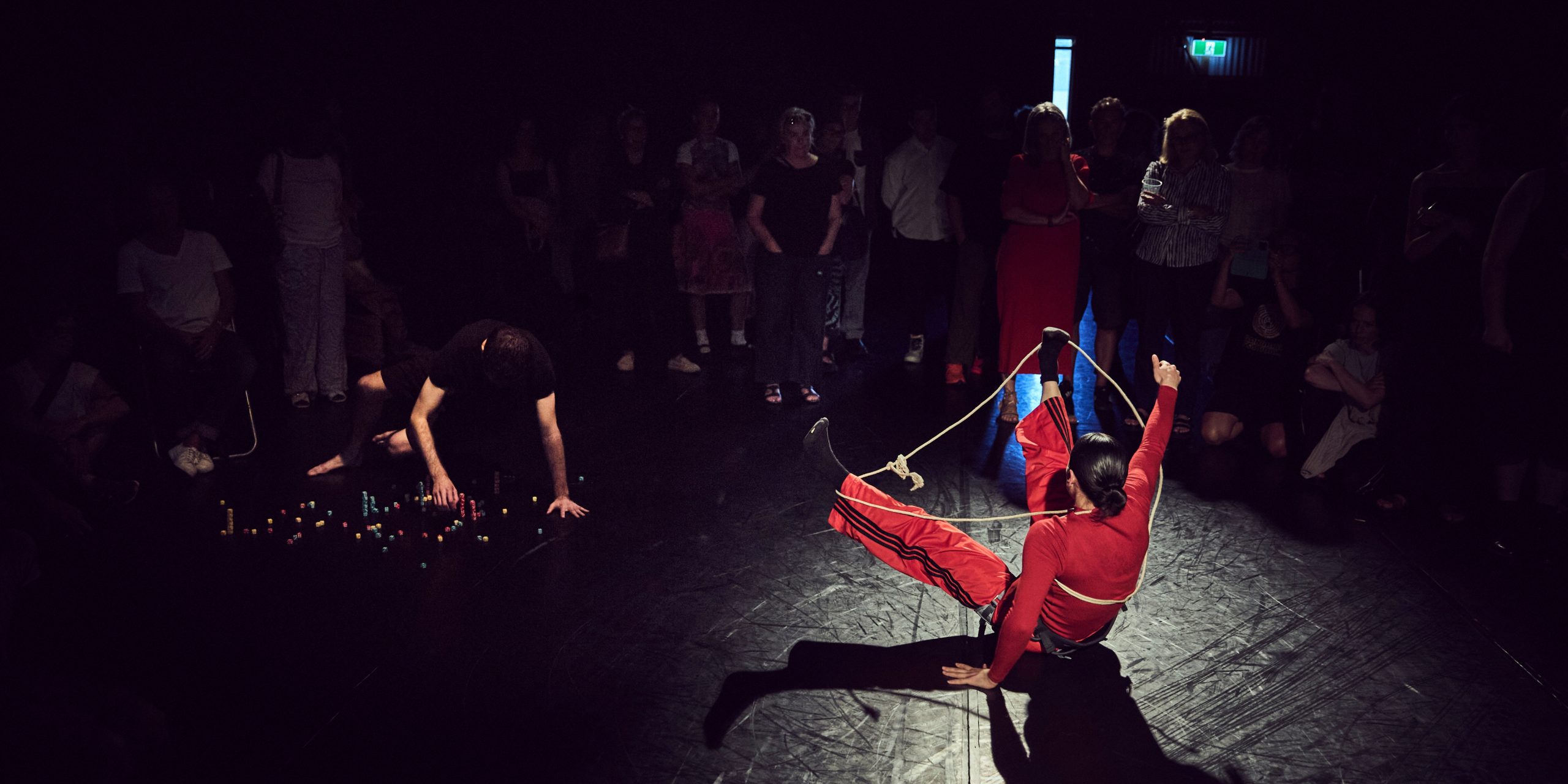 An image taken at nighttime outside The Drill Hall on lawn beside the harbour. A person stands holding onto a large yellow ladder. Another person is captured in flight diving off the ladder into an inflatable baby pool.