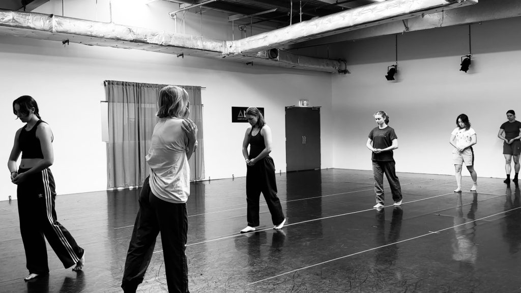6 dancers pictured in a studio positioned on their knees on the floor with their arms crossed.