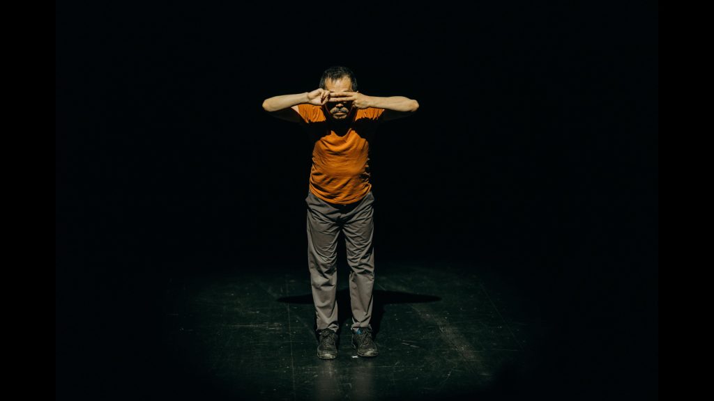 A person is standing at the centre of dark performance space. They are leaning slightly forward towards the audience. Their arms are bent and fingers of both hands meeting in front of their eyes like two parallel train tracks.