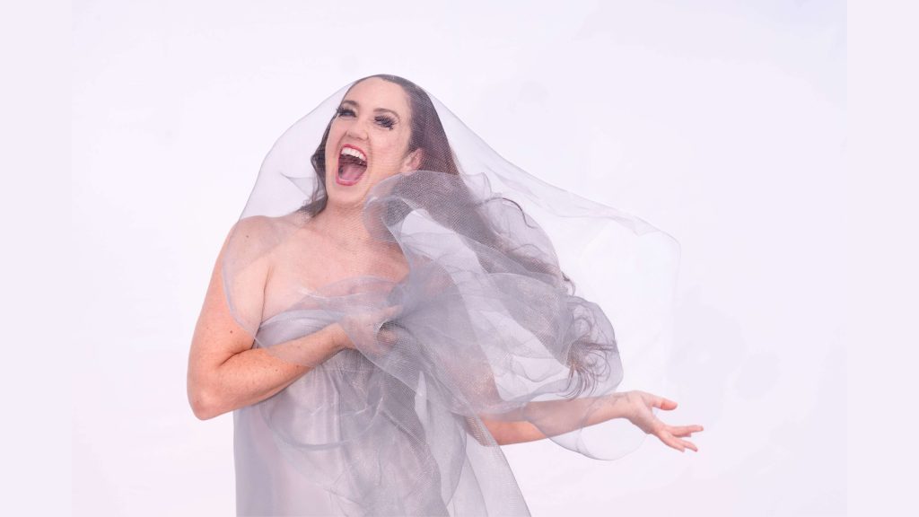 A photograph of a woman wearing silver sheer fabric. She has a wonderfully expressive face with her mouth wide open in a large smile.