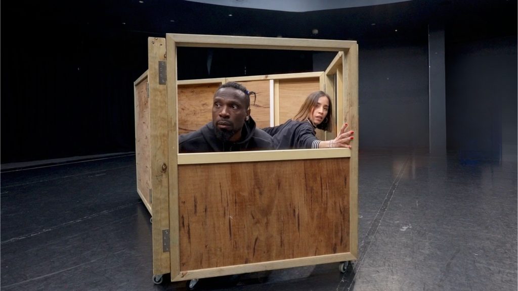 A photograph of two people sitting inside a large timber packing box.