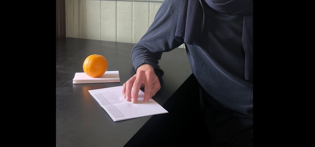 A person dressed in navy has their hand on an open booklet. An orange sits on top of a stack of paper. They are reading. 