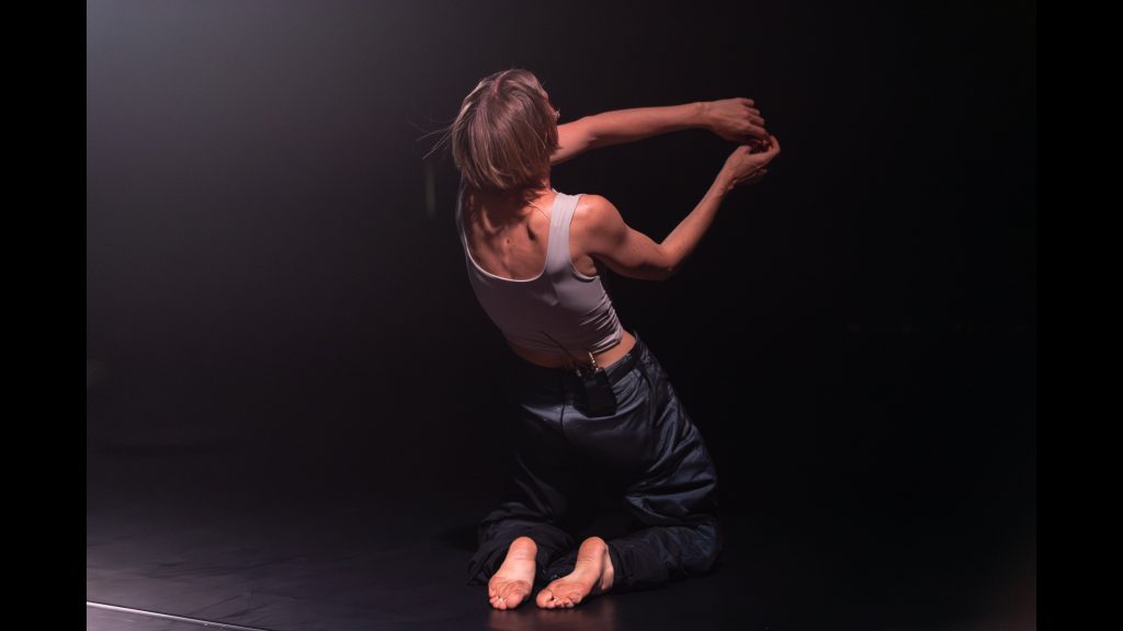 A performer is kneeling on floor, leaning back, with arms forward and fingers touching loosely. Photo is taken from behind the performer and face is not visible.