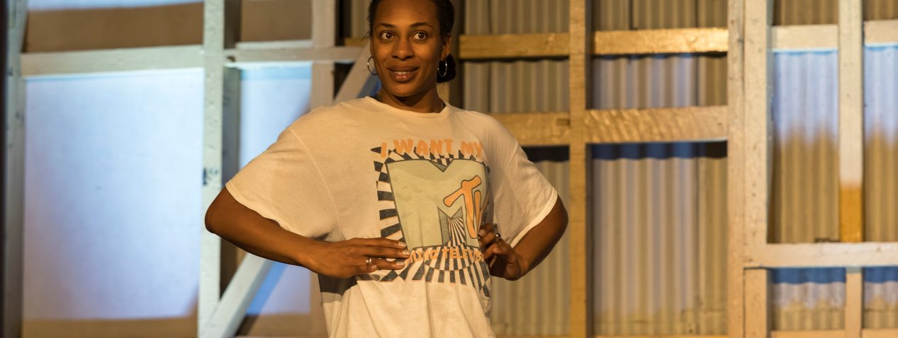 Photograph of a person performing. They have their hands on their waster and are smiling.