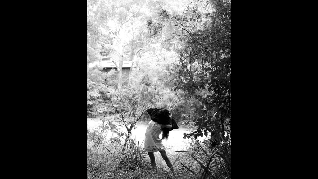 Black and White photograph of trees in a park. In teh foreground, there is a person wearing a dress and their face and head are covered with a fabric.