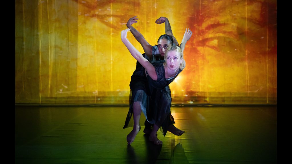 An image of two dancers performing in Sue Healey's work called AFTERWORLD. Both dancers are leaning forward with their arms reaching back behing them, wrists folded inwards and faces visible with gaze up and outwards. Behind the performers, there is a large projection of a person's eyes.
