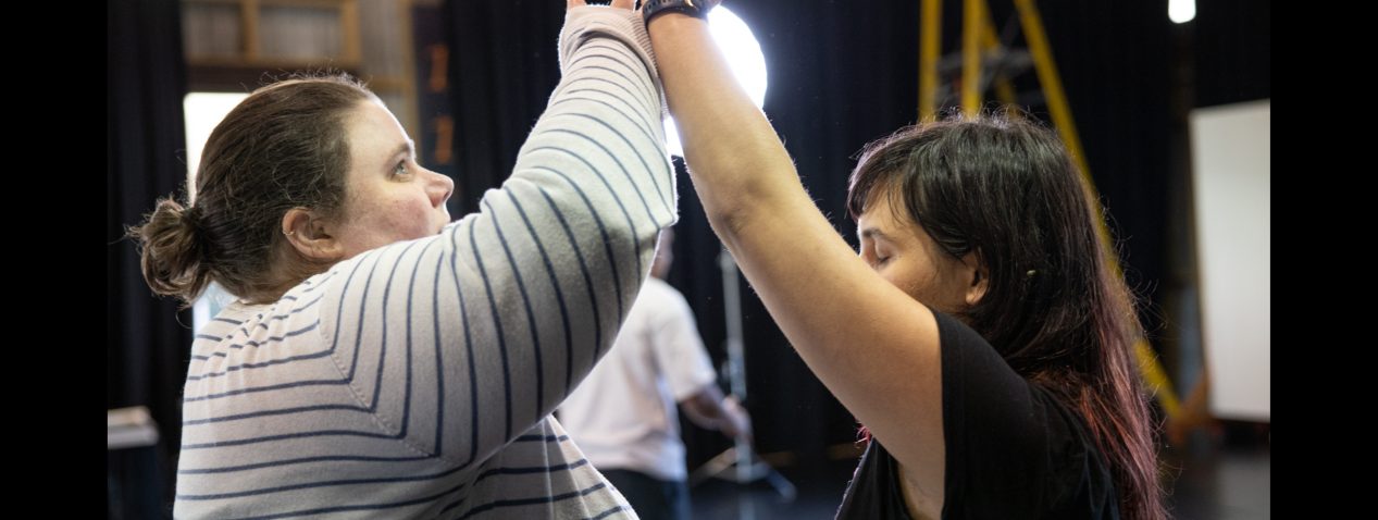 Image of two people facing each other. They are mirroring each other through their movement, both with an arm reaching up high and in contact with each other's wrist.