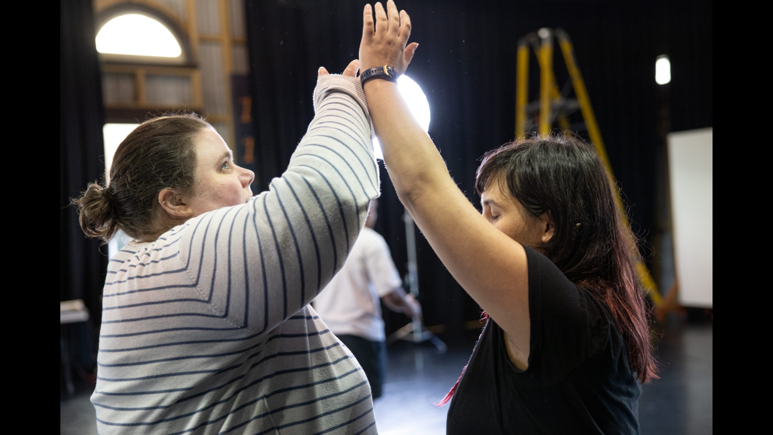 Image of two people facing each other. They are mirroring each other through their movement, both with an arm reaching up high and in contact with each other's wrist.