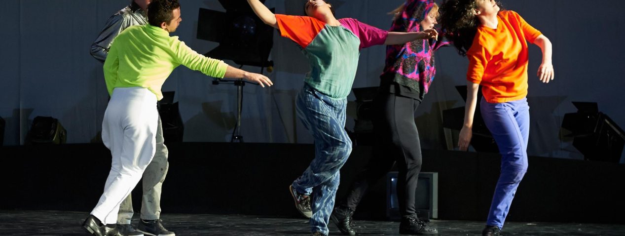 An image of a group of five people colourfully dressed, dancing on a stage. The person in the centre of the group has their upper back arched, head back, arms outstretched, and one foot resting on the other lower leg, almost like a bird.