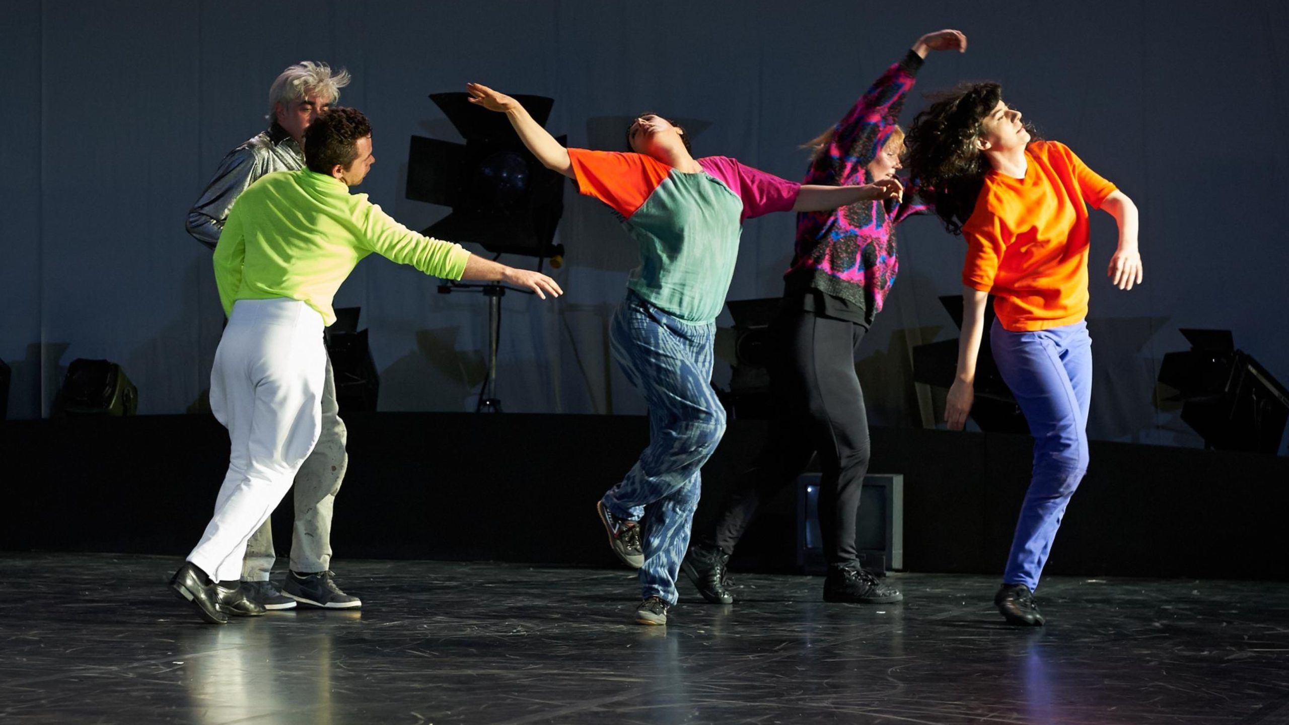 An image of a group of five people colourfully dressed, dancing on a stage. The person in the centre of the group has their upper back arched, head back, arms outstretched, and one foot resting on the other lower leg, almost like a bird.