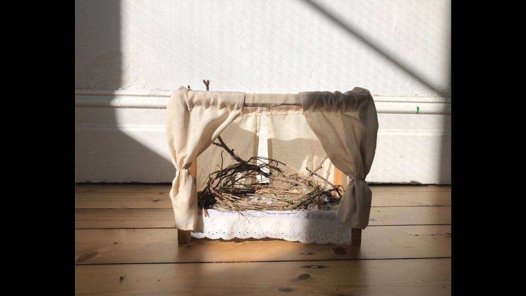 The image shows an artwork titled ‘Nest’ created during Iris Elgar’s Front Studio Residency at Temperance Hall. A dollhouse four-poster bed is occupied by a nest constructed in the fashion of a House Sparrow’s. It is made up of twigs, dried grass, fabric offcuts, leaves, dust, feathers, and hair. 
