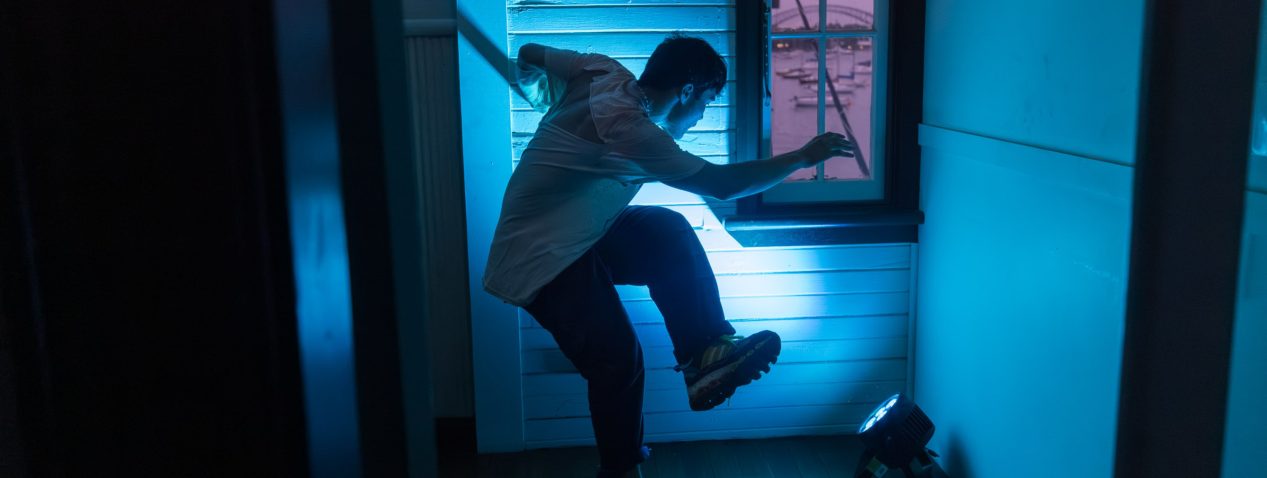 Photograph of a dancer balancing on the edge of one foot with other leg lifted up. The room is lit blue and it contrasts against the view out a small window with a pink sunset.