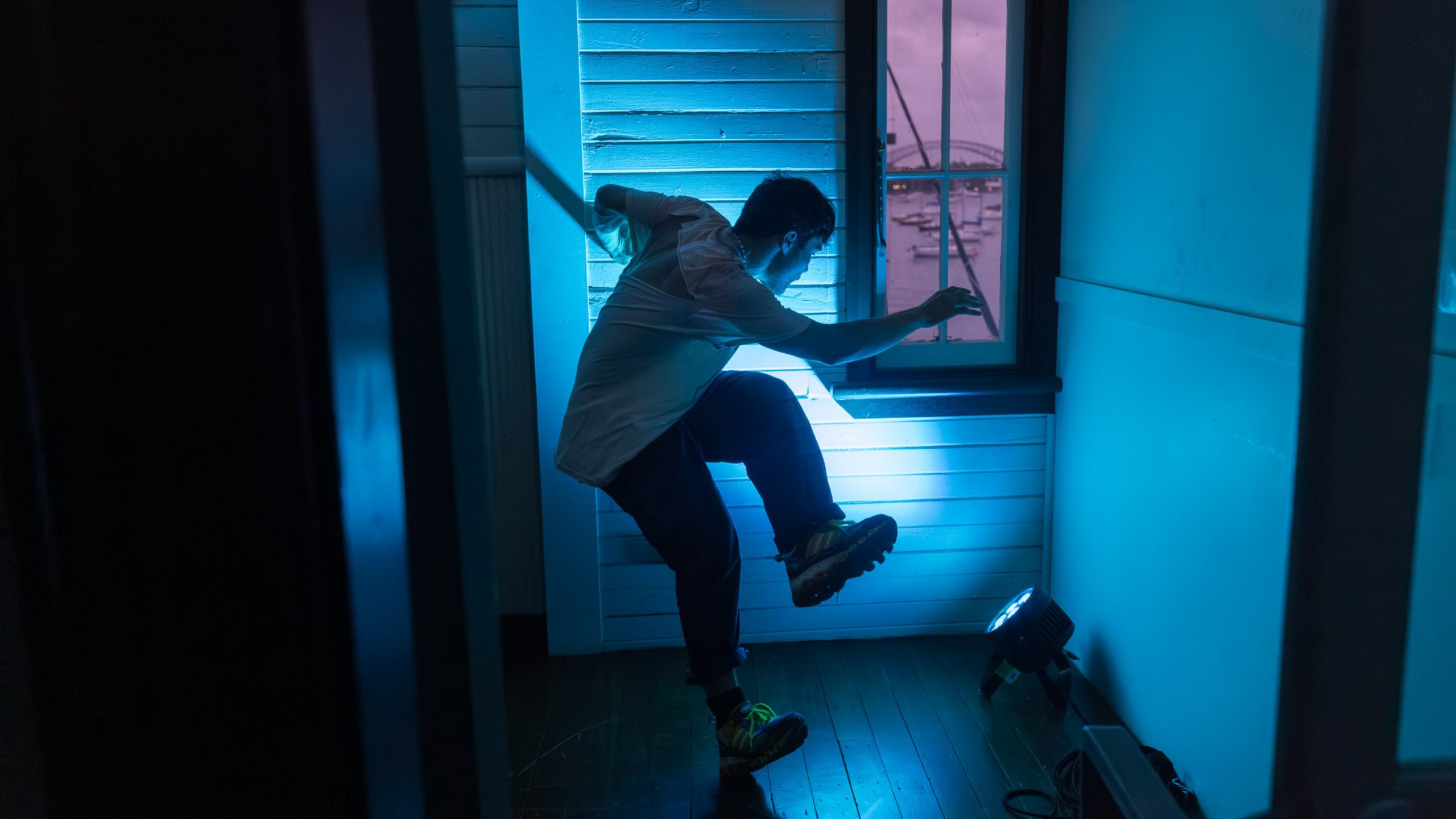Photograph of a dancer balancing on the edge of one foot with other leg lifted up. The room is lit blue and it contrasts against the view out a small window with a pink sunset.