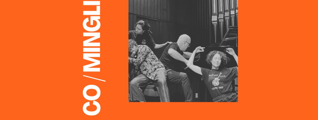 A photo of 5 performers, they are all in physical contact with with at least 2 other performers. One is holding up a microphone up to their lips and another is playing a piano. There are 2 signs saying "Caution, Wet Floor".