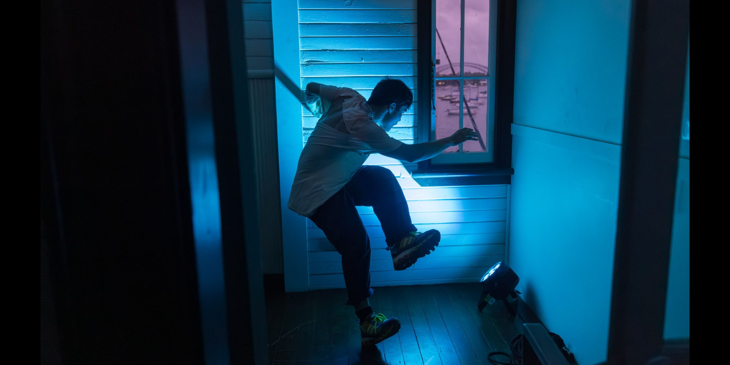 Photograph of a dancer balancing on the edge of one foot with other leg lifted up. The room is lit blue and it contrasts against the view out a small window with a pink sunset.