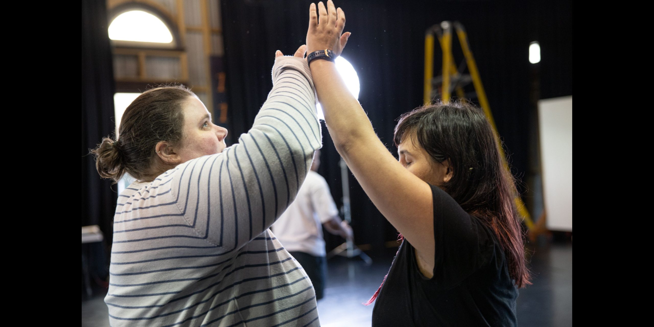 Image of two people facing each other. They are mirroring each other through their movement, both with an arm reaching up high and in contact with each other’s wrist.