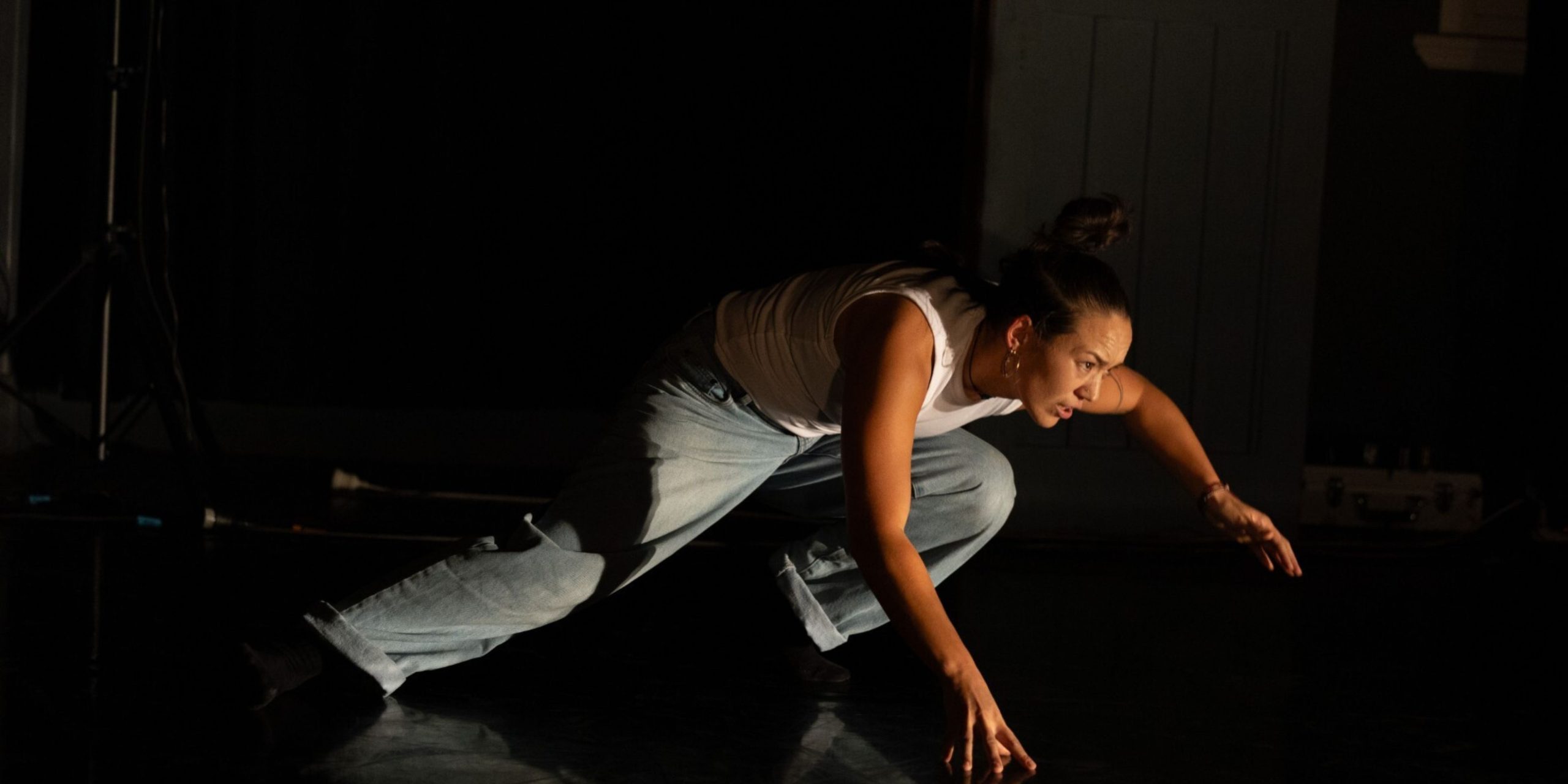 A lively group of dancers moves expressively across a large dance studio. At the centre, a dancer in pink pants and a white top leads with joyful energy, arms open wide. The other dancers follow in rhythmic, expansive gestures, filling the space with motion and connection.