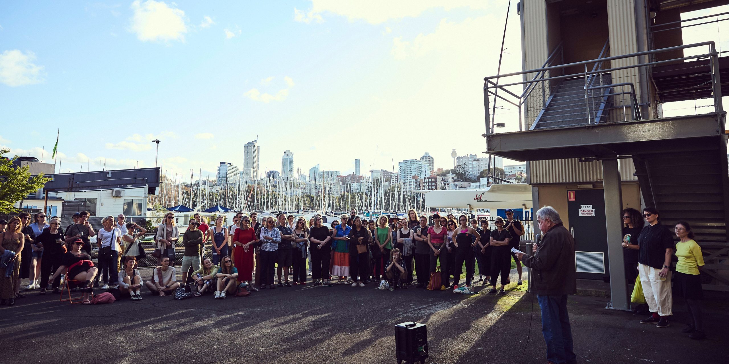 A lively group of dancers moves expressively across a large dance studio. At the centre, a dancer in pink pants and a white top leads with joyful energy, arms open wide. The other dancers follow in rhythmic, expansive gestures, filling the space with motion and connection.