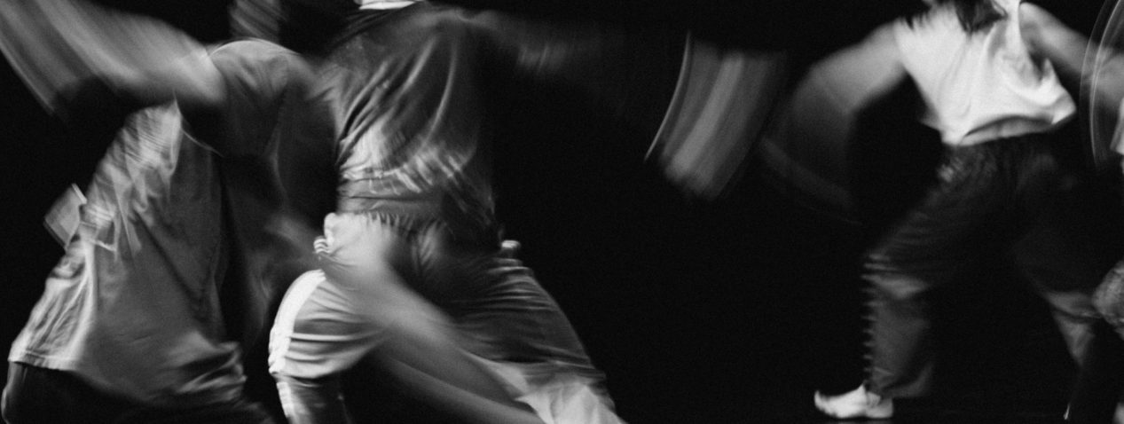 A black and white photograph of three dancers captured in flight, slightly blurred with arms out, weight on left foot and right leg stretched out behind the body.