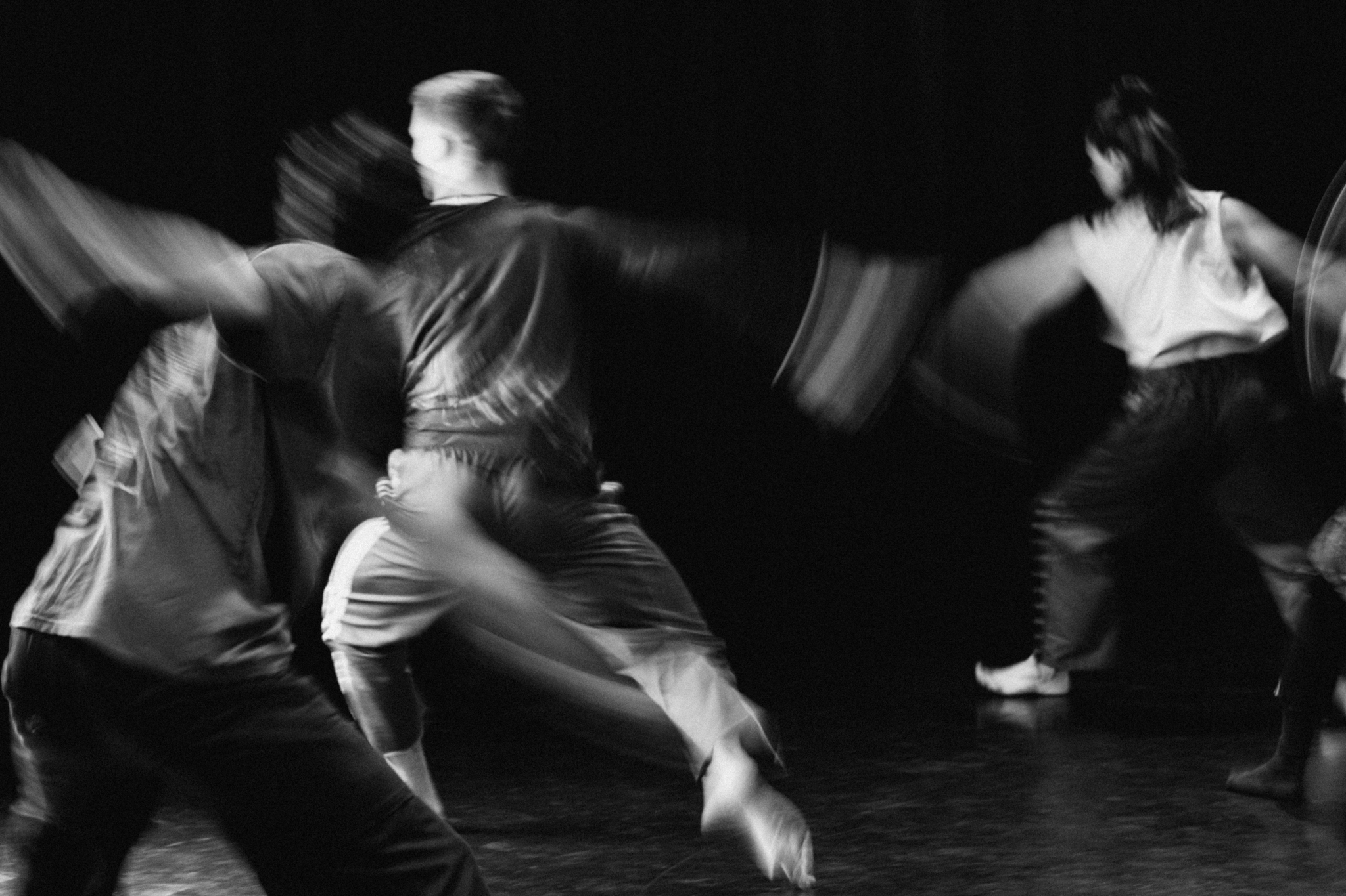 A black and white photograph of three dancers captured in flight, slightly blurred with arms out, weight on left foot and right leg stretched out behind the body.