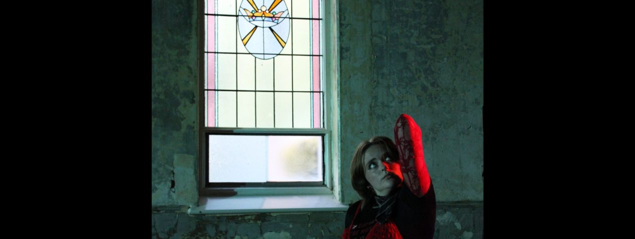 A photo of a person standing inside by a stained glass window. They are looking up and their arm is folded with the elbow reaching upwards too.