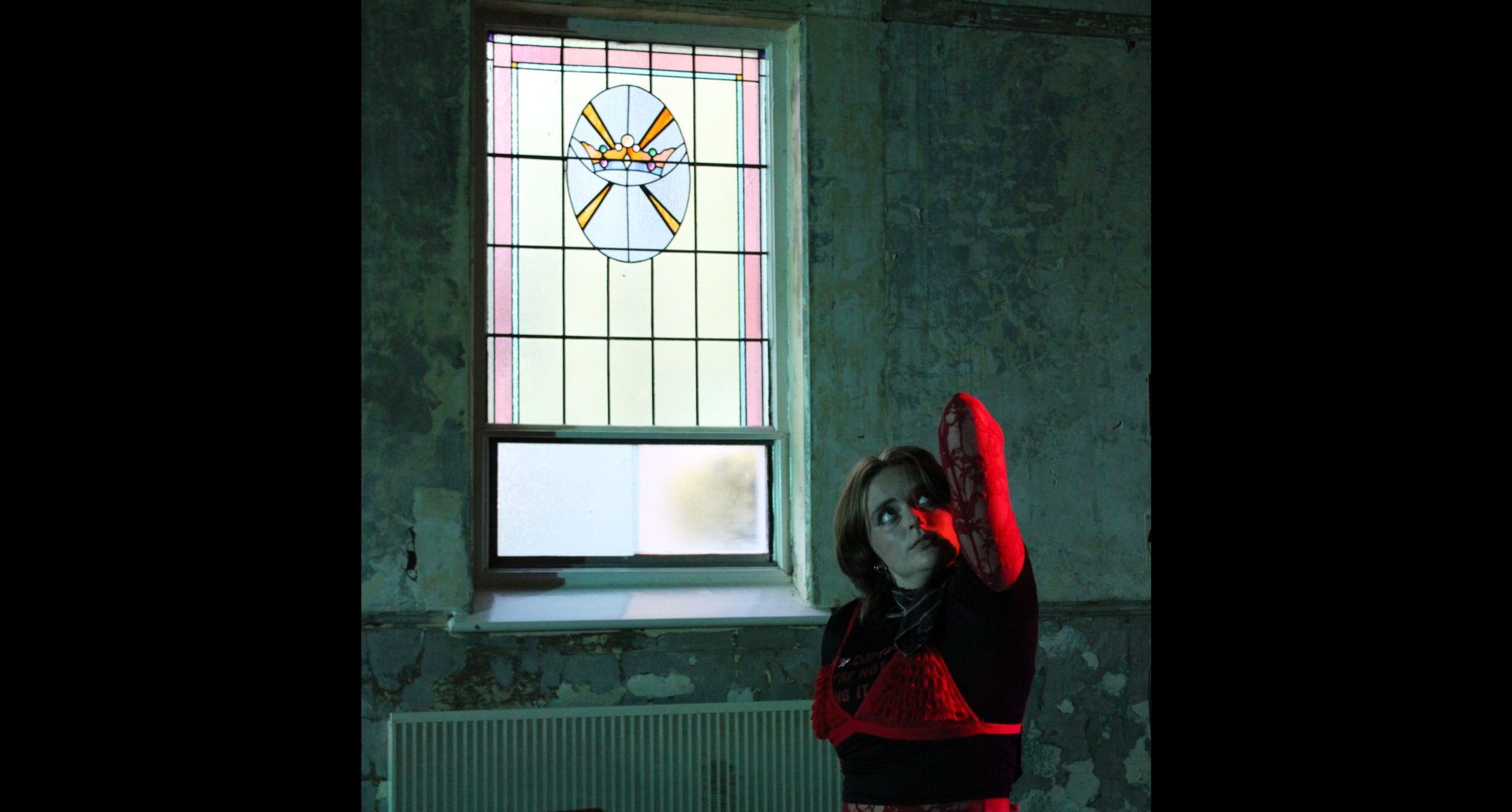 A photo of a person standing inside by a stained glass window. They are looking up and their arm is folded with the elbow reaching upwards too.