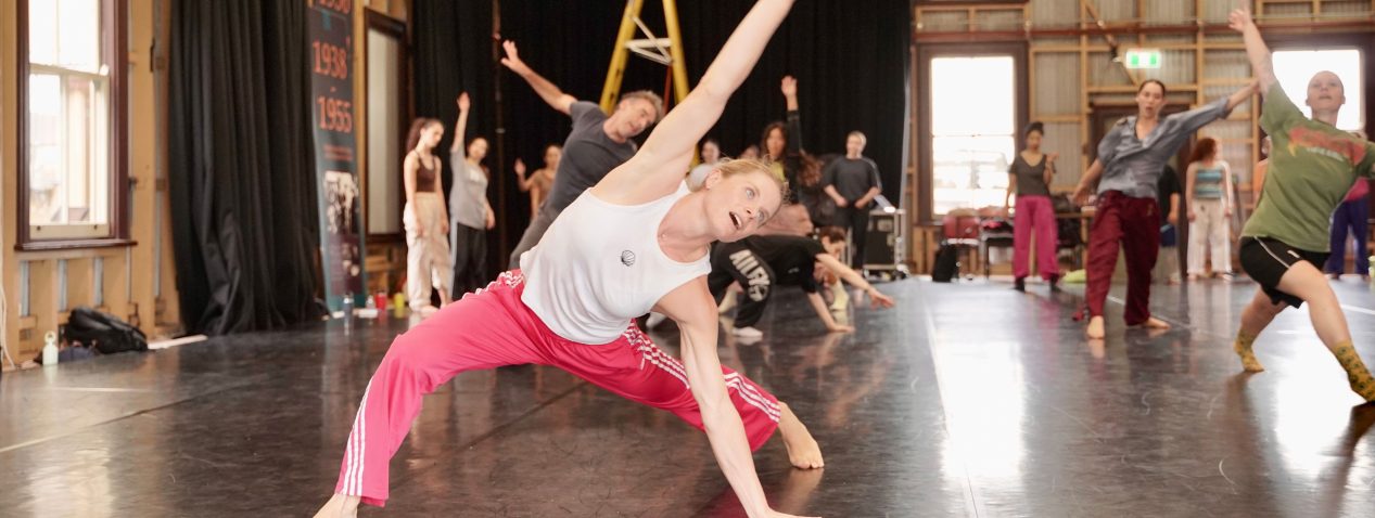A group of adults practices dance in a large studio. In the foreground, a woman wearing a white sleeveless top and bright pink pants stretches sideways with one hand on the floor and one arm extended overhead. Other dancers behind her mirror the movement, with their arms raised or bodies leaning to the side. The studio has a polished dark floor, high exposed beams, large windows, and black curtains.