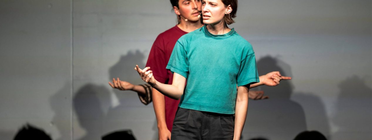 A photograph of four performers standing in a line behind each other. The person in front is speak with their head turned to the right and gesturing with their right hand. The person behind them is speaking with their head turned to the left and gesturing with their left hand. The two people behind them are hidden all except for their hands.