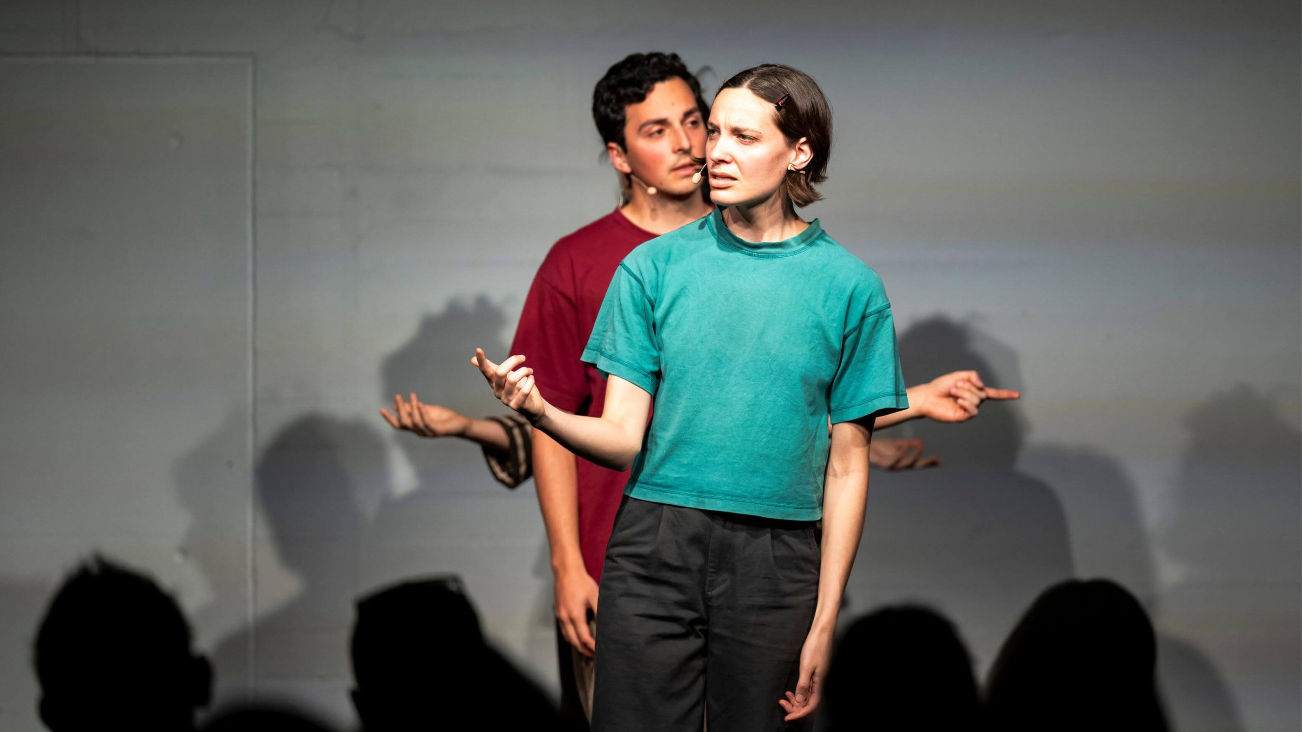 A photograph of four performers standing in a line behind each other. The person in front is speak with their head turned to the right and gesturing with their right hand. The person behind them is speaking with their head turned to the left and gesturing with their left hand. The two people behind them are hidden all except for their hands.