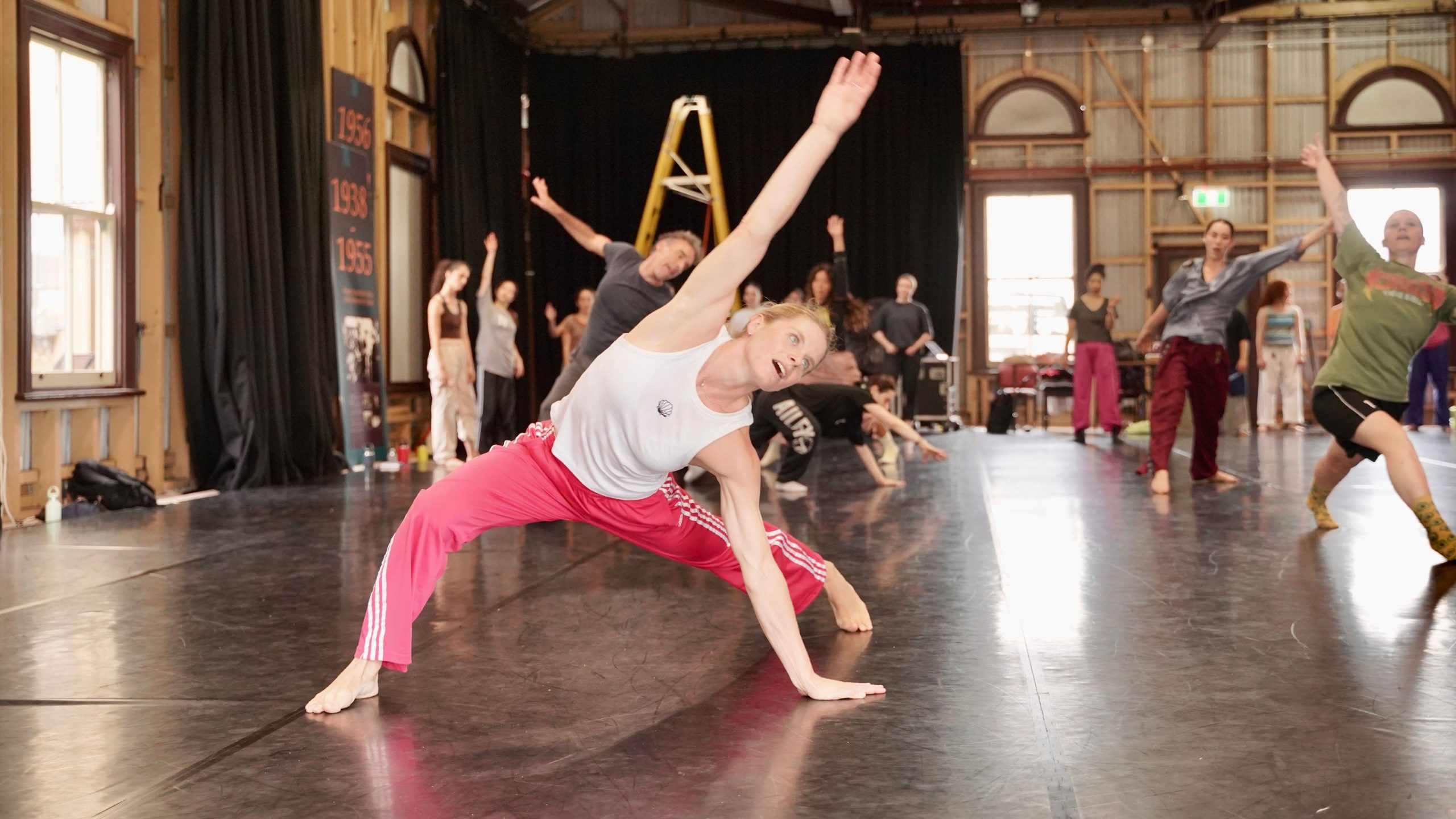 A group of adults practices dance in a large studio. In the foreground, a woman wearing a white sleeveless top and bright pink pants stretches sideways with one hand on the floor and one arm extended overhead. Other dancers behind her mirror the movement, with their arms raised or bodies leaning to the side. The studio has a polished dark floor, high exposed beams, large windows, and black curtains.