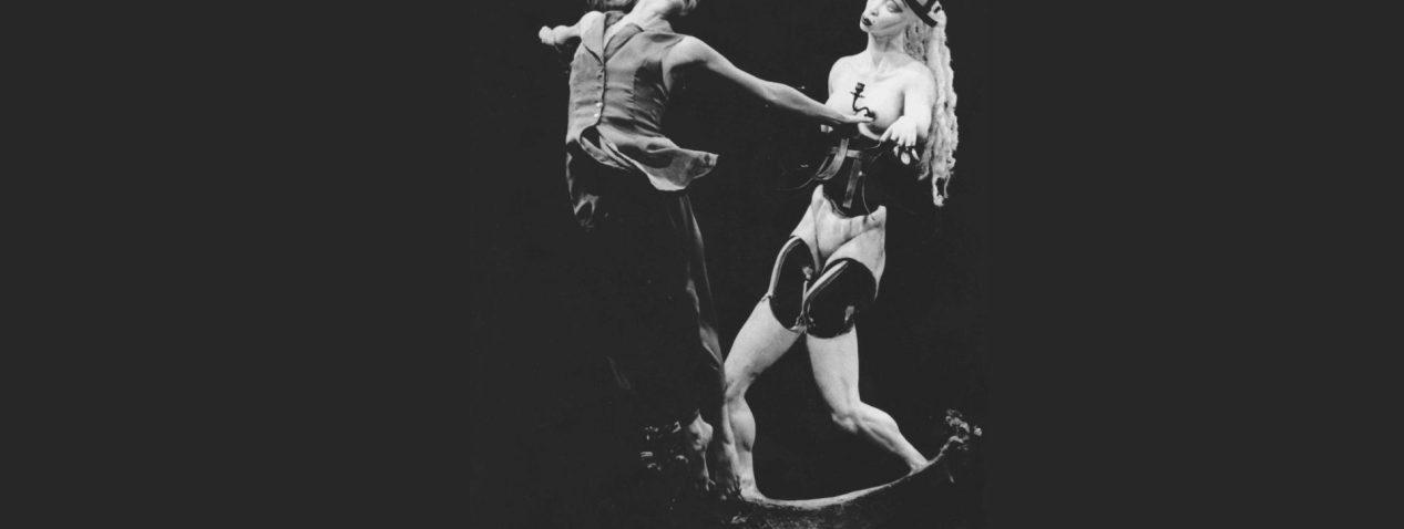 An image of a dancer captured in flight, jumping with her back arched, eyes closed and arms stretching behind her. Beside the dancer is a figure with a face and eyes gazing towards the dancer. The figure is a readymade object constructed from mannequin parts and other materials.